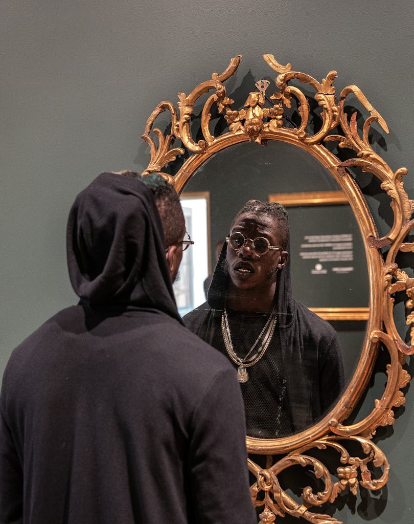 Stylish man wearing a hoodie and sunglasses looks into an ornate mirror indoors.